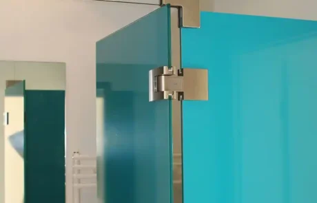 Nahaufnahme einer gläsernen Duschtür mit Edelstahlscharnieren, einer türkisfarbenen Glasscheibe und einem Teil eines Badezimmers im Hintergrund.