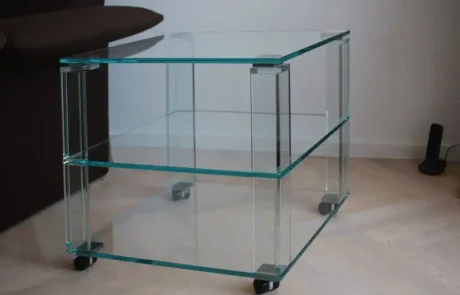 Ein Beistelltisch aus Klarglas mit zwei Ablagen und vier Rollen, der auf einem hellen Holzboden neben einem dunkelbraunen Sofa steht.