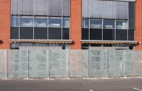 Ein Backsteingebäude mit großen Fenstern und Metalljalousien hat einen dekorativen Milchglaszaun mit einem Blattmuster vor sich.