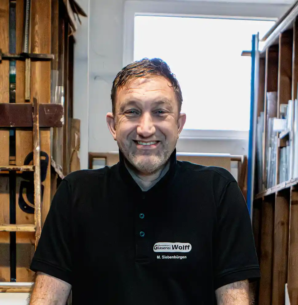 Buntentors_Glaserei_Wolff_Bremen_Siebenbuergen_Kontakt Ein Mann in einem schwarzen Polohemd mit dem Namensschild "Glaserei Wolff" steht in einem Innenraum und lächelt in die Kamera, während im Hintergrund Regale und Holzrahmen zu sehen sind.